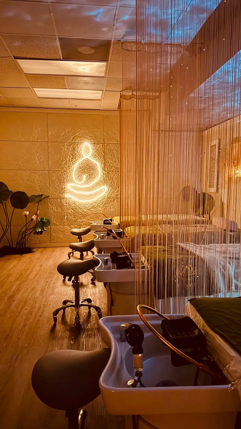 The Zenvie head spa room — white shampoo basins and stools beside the illuminated lotus sign, with lotus plants and beaded-curtain dividers in warm light